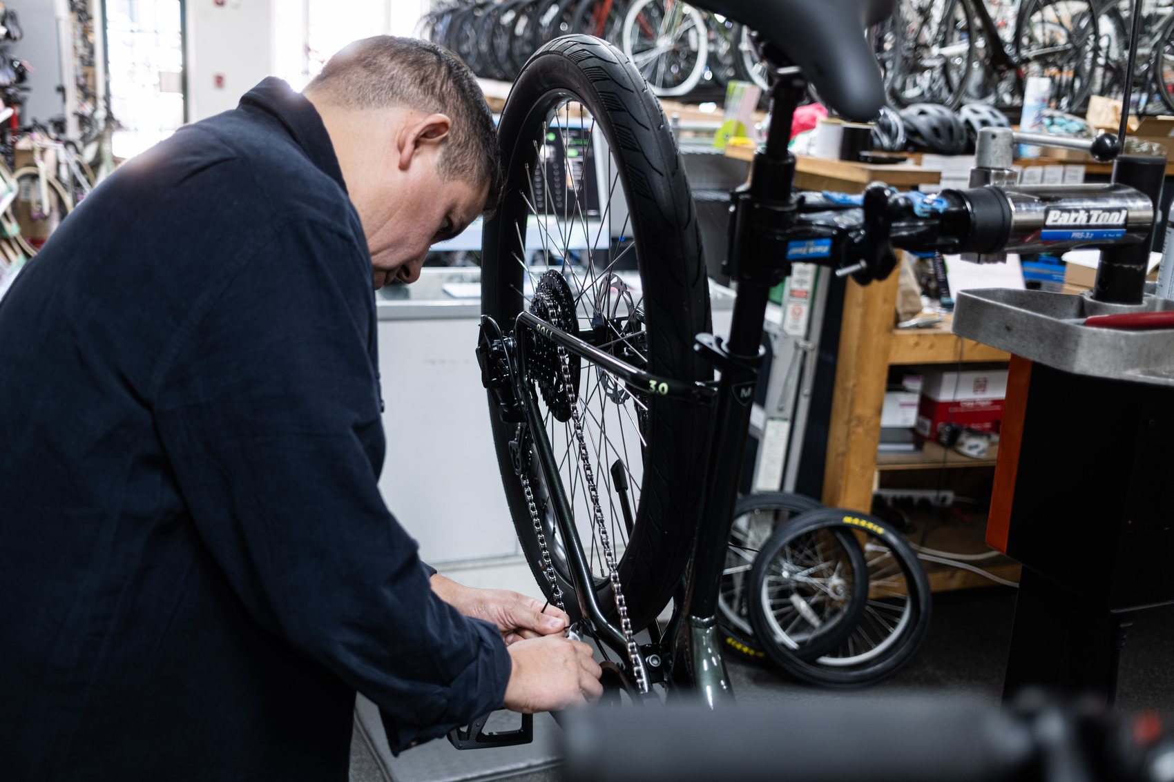 A mechanic leaning over a bike in a Park Tool repair stand, adjusting the rear drivetrain, with the shop's bike racks behind him