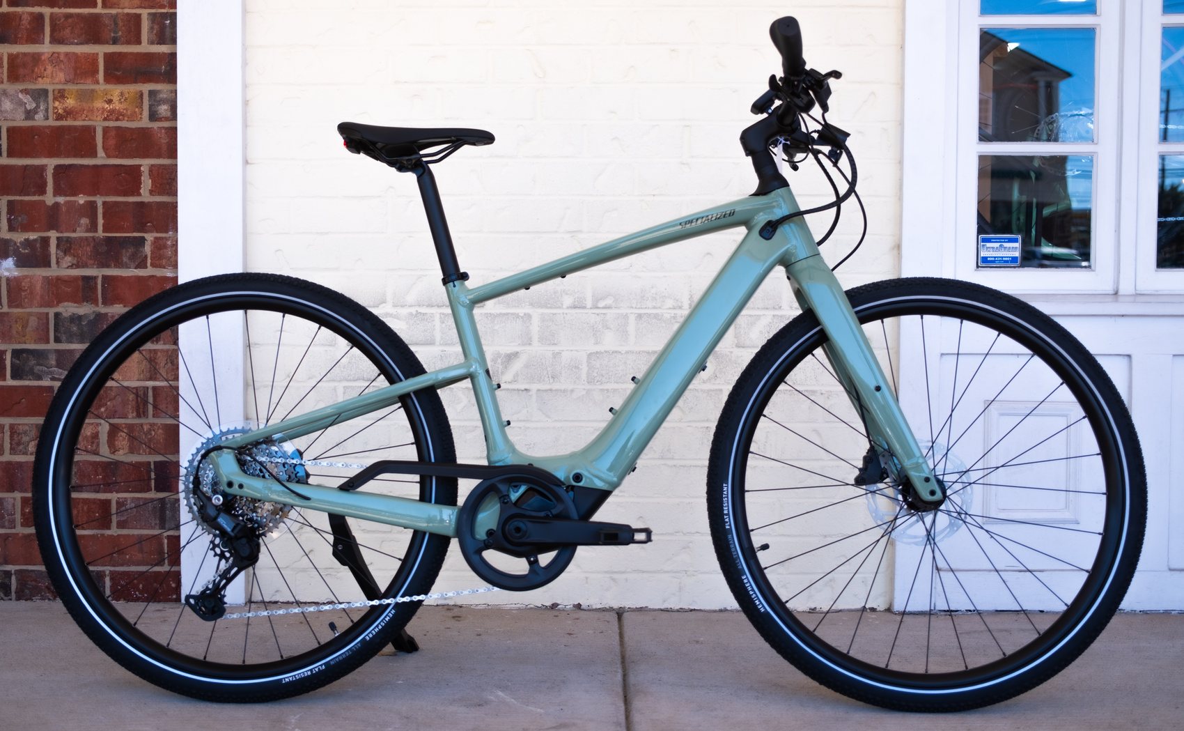 Sage green Specialized Turbo Vado SL e-bike parked on the sidewalk outside the shop, seen from the side against a cream brick wall